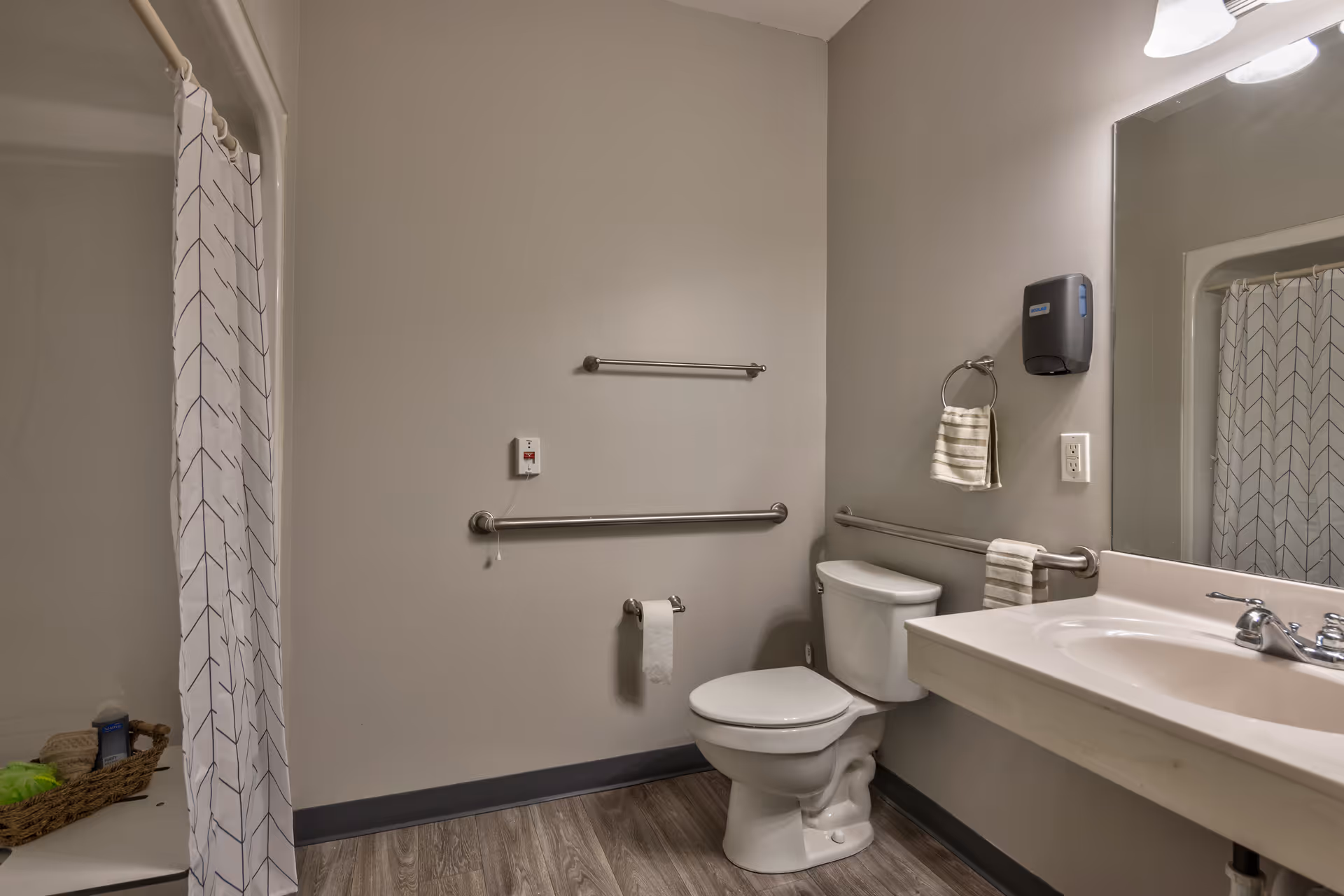 A clean and accessible bathroom with a toilet, a sink with a faucet, a large mirror, and a shower area with a white curtain featuring a simple arrow pattern. The bathroom has grab bars near the toilet and sink, a towel ring with a striped hand towel, a soap dispenser mounted on the wall, and a basket with toiletries in the shower area. The floor has wood-like vinyl flooring and the walls are painted light gray.
