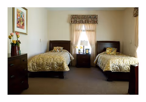 A bedroom with two twin beds, each with a patterned gold bedspread and decorative pillows. Between the beds is a nightstand with a lamp, a framed photo, and a small vase of flowers. The window behind the nightstand has sheer curtains with a floral valance. To the left is a dark wooden dresser with a vase of flowers and a framed picture hanging above it. The room has beige walls and carpeted floor.