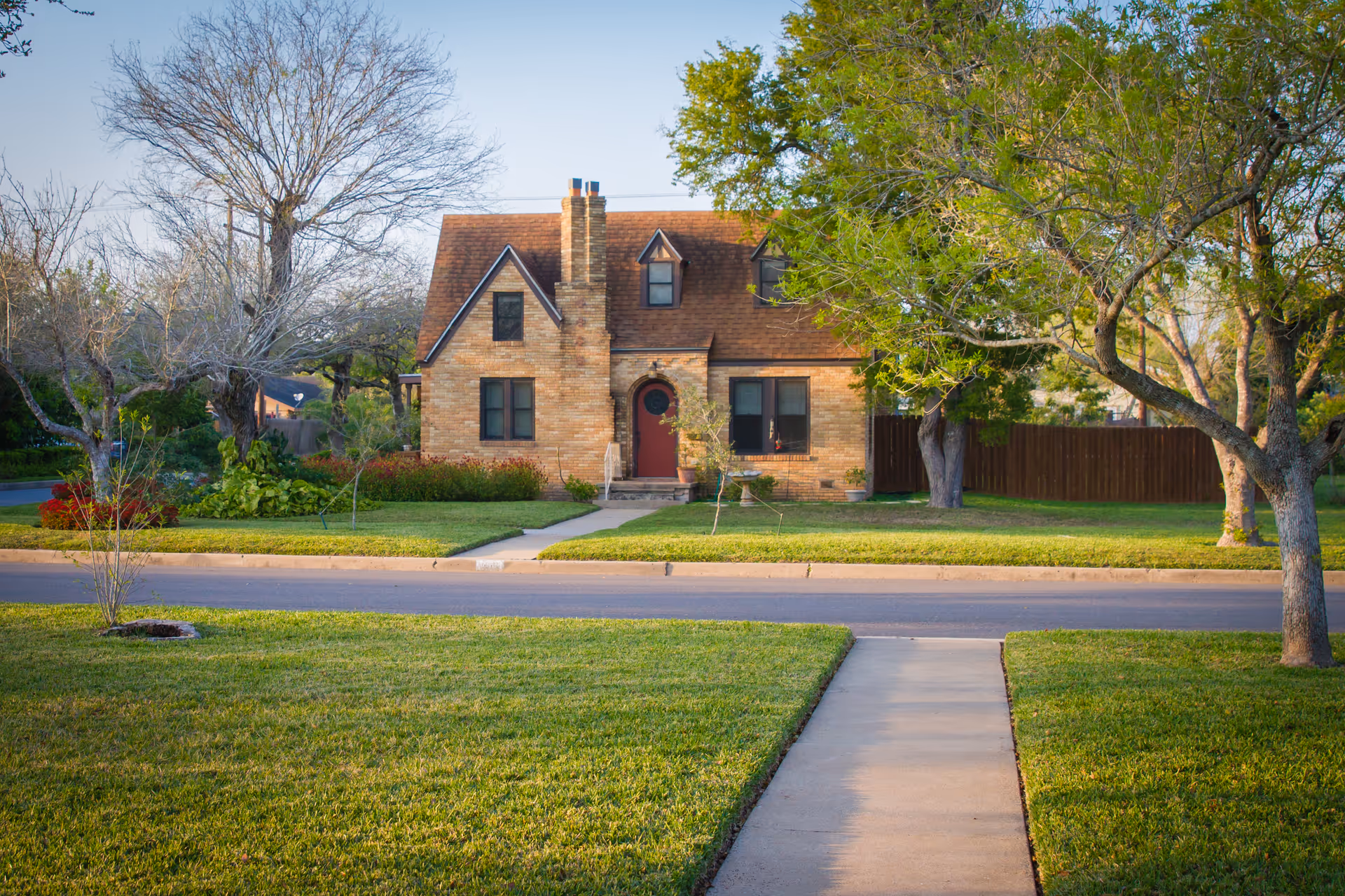 A charming brick house with a steep roof and chimney, surrounded by a well-maintained lawn and trees, viewed from across the street with a sidewalk leading up to the front door.