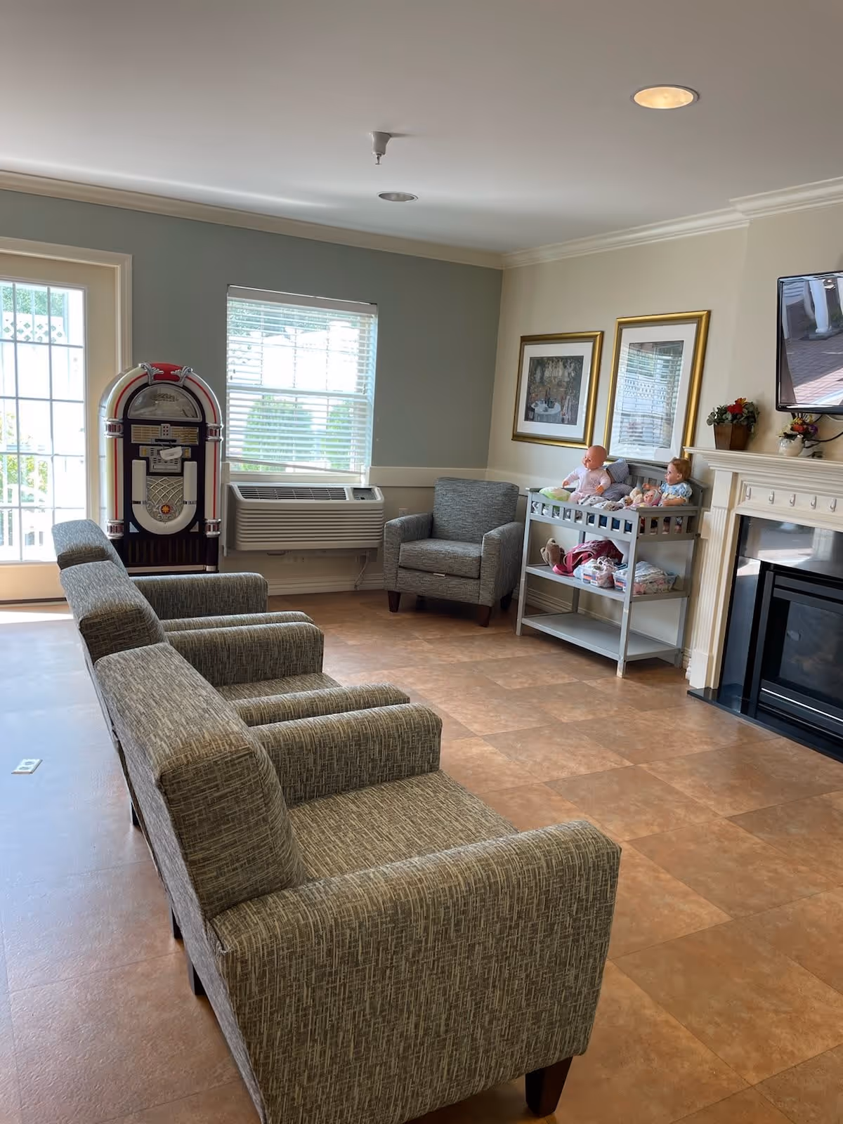 A cozy living room area with three gray upholstered armchairs arranged in a row facing a fireplace. There is a single gray armchair near a window with blinds, and a vintage-style jukebox next to a glass door. A small table with dolls and framed pictures on the wall is beside the fireplace. The room has a tiled floor and neutral-colored walls.