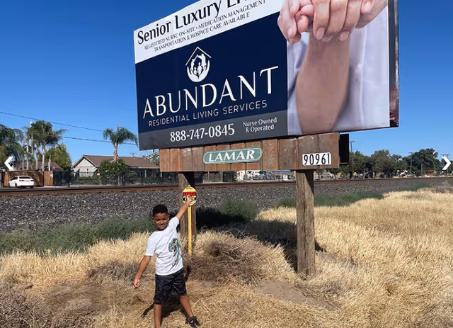 A young boy stands in front of a large billboard advertising Abundant Residential Living Services, a senior luxury living facility. The billboard includes contact information and mentions services like registered nurse on-site, medication management, transportation, and hospice care. The setting is outdoors near a railroad track with dry grass and some houses and palm trees in the background.