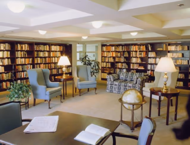 Library-style common room with bookshelves lining the walls, upholstered armchairs, side tables with lamps, and a globe.