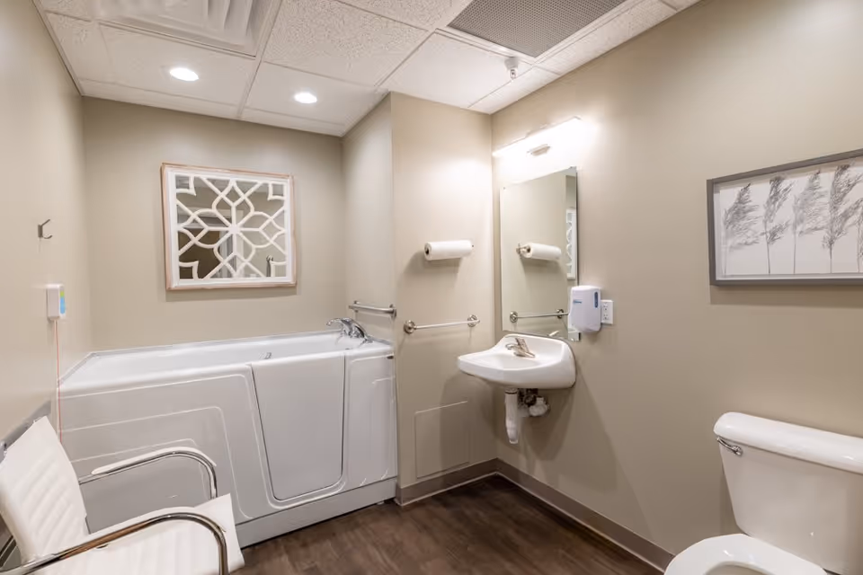 A clean, accessible bathroom with a walk-in bathtub, a white sink with a mirror above it, a toilet, a white chair with metal armrests, and beige walls decorated with a framed mirror and a framed picture of trees.