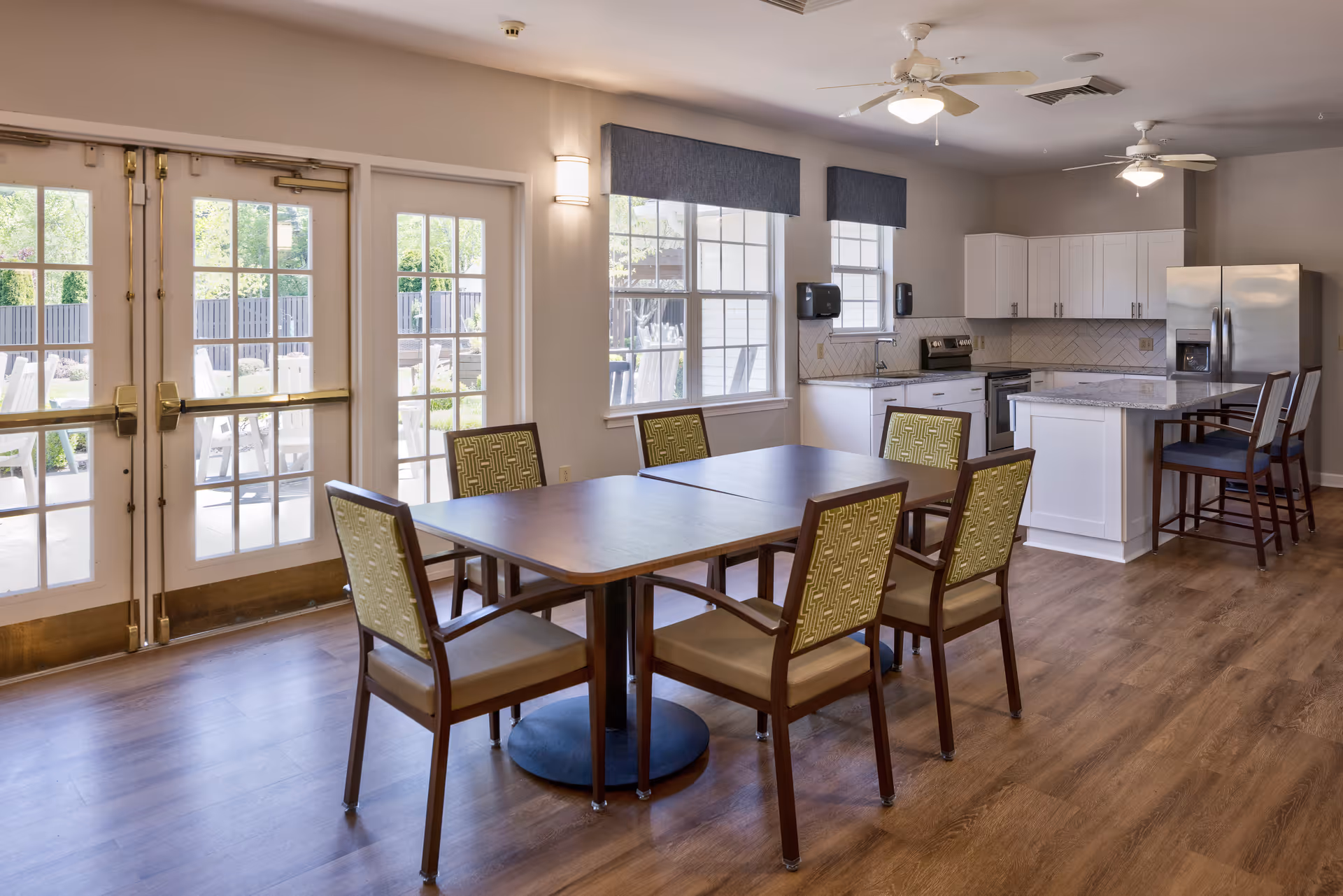 A bright and spacious dining area with a wooden table surrounded by six chairs with patterned upholstery. The room features large windows with blue valances, double glass doors leading outside, and a modern kitchen in the background with white cabinets, stainless steel appliances, and a kitchen island with two bar stools. The floor is wood, and ceiling fans with lights are mounted on the ceiling.