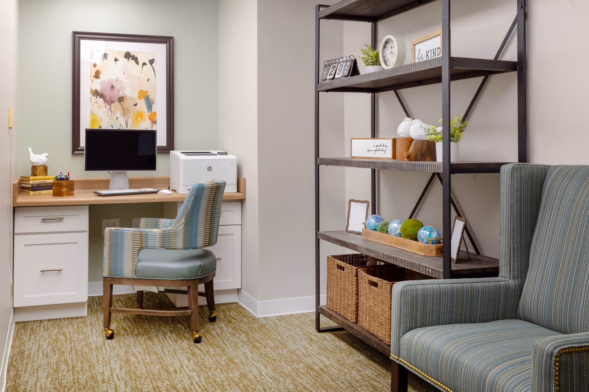 A cozy office corner in a senior living facility featuring a wooden desk with a computer monitor, keyboard, mouse, and printer. A patterned upholstered chair with wheels is positioned at the desk. To the right, there is a tall metal and wood shelving unit decorated with plants, framed signs, decorative balls, and wicker baskets. A striped upholstered armchair is partially visible in the foreground. The room has light-colored walls and carpeted flooring.