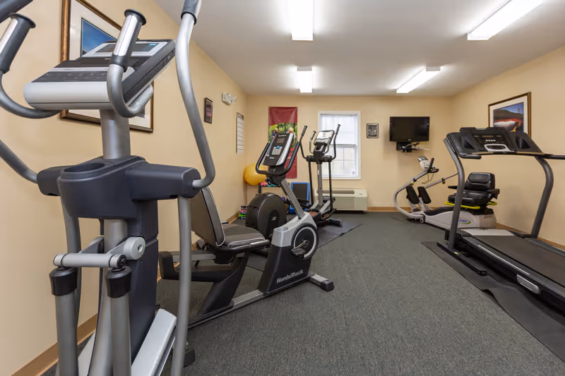 A small fitness room with exercise equipment including a recumbent bike, elliptical machine, treadmill, and rowing machine. The room has beige walls, a window with white blinds, framed pictures on the walls, and fluorescent ceiling lights.