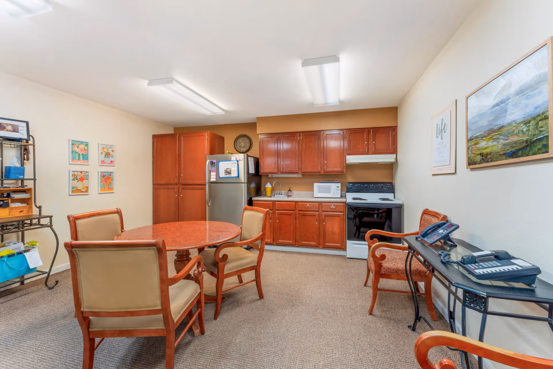 A communal kitchen and dining area with wooden cabinets, a stainless steel refrigerator, a white stove, and a microwave on the countertop. There is a round wooden table with four upholstered chairs in the center of the room. On the right side, there is a glass-top table with two telephones and two chairs. The walls are decorated with framed artwork and a motivational quote. The floor is carpeted.