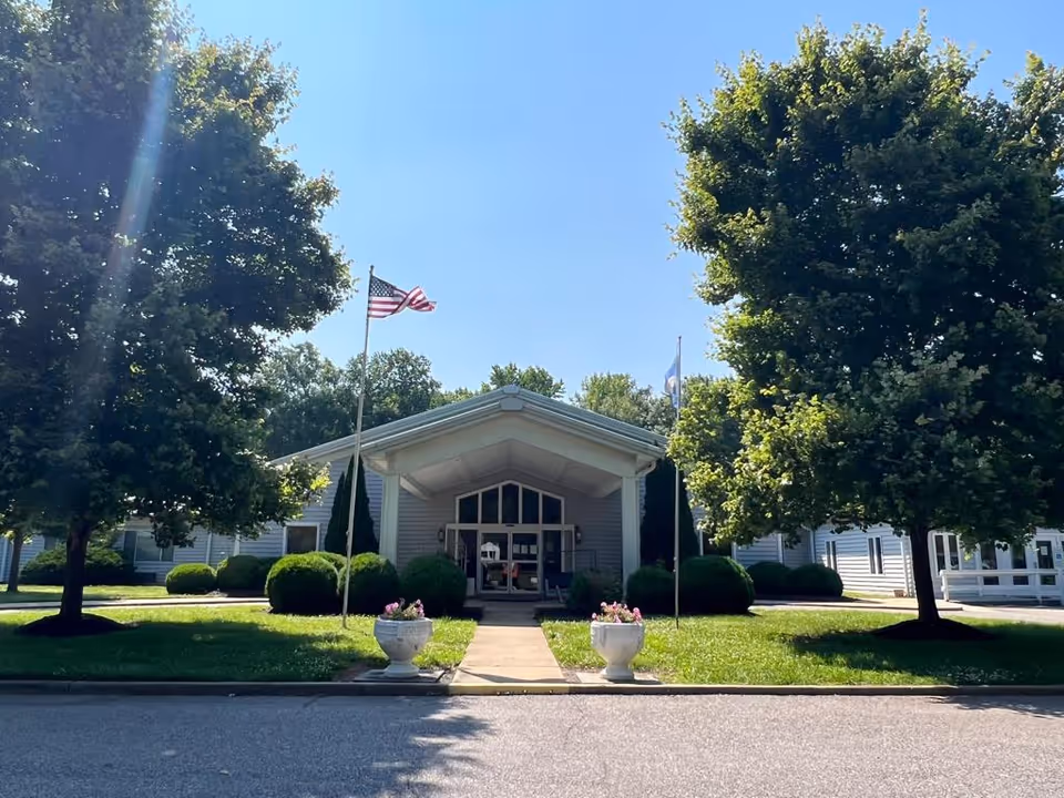 Front exterior view of Amelia Rehabilitation and Healthcare Center building with a covered entrance, two flagpoles with American and another flag, green bushes, two large trees, and flower pots on either side of the walkway leading to the entrance.