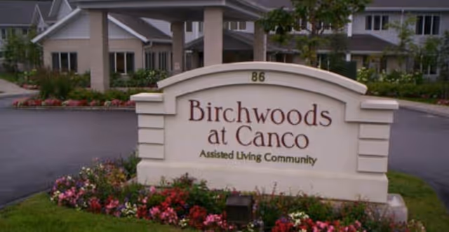 Entrance sign for Birchwoods at Canco Assisted Living Community with a landscaped flower bed in front and residential buildings in the background.
