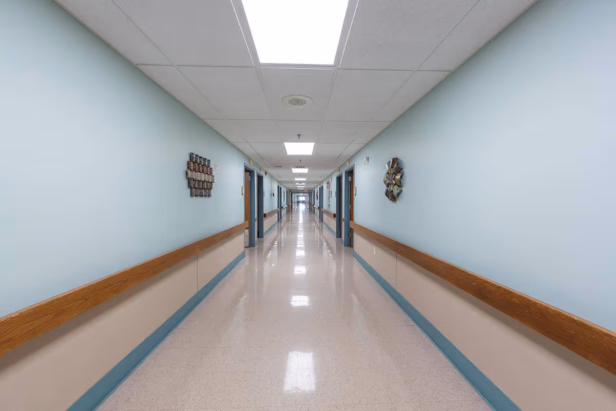 A long, clean hallway in a healthcare facility with light blue walls, beige lower wall panels, wooden handrails on both sides, and several doorways along the corridor. The ceiling has recessed lighting panels and the floor is shiny and reflective.