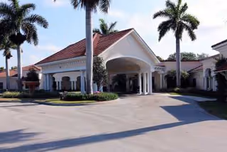 Exterior view of a single-story building with a covered entrance supported by white columns, surrounded by palm trees and landscaping under a clear sky.