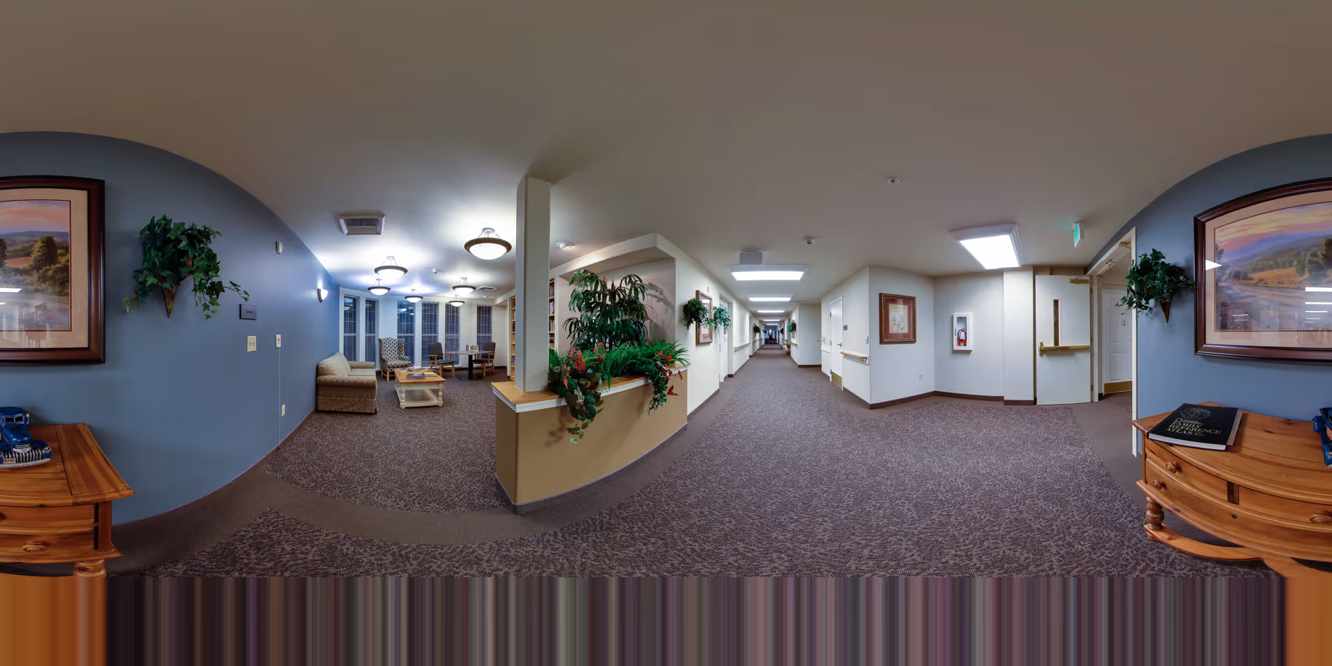 Long carpeted interior corridor of a senior living facility with a seating/lounge area, potted plants, framed artwork and ceiling lights.