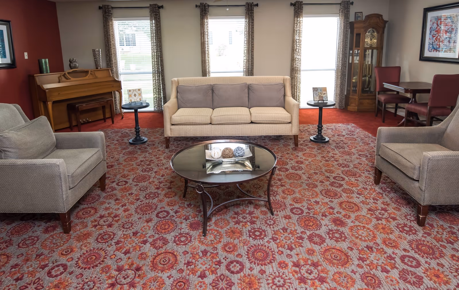 A cozy living room with a patterned red and orange carpet, a beige sofa with three gray cushions, two matching armchairs, a round glass coffee table with decorative items, two small round side tables, a wooden piano against a red accent wall, a tall wooden grandfather clock, and a small dining table with four chairs near a window with patterned curtains.