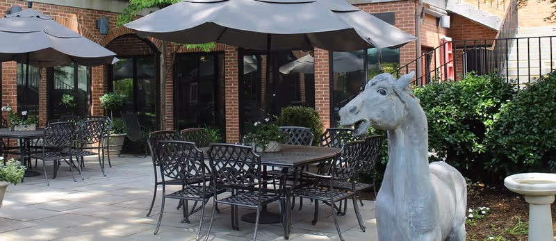 Outdoor courtyard with metal patio tables and umbrellas in front of a brick building and a stone horse statue.