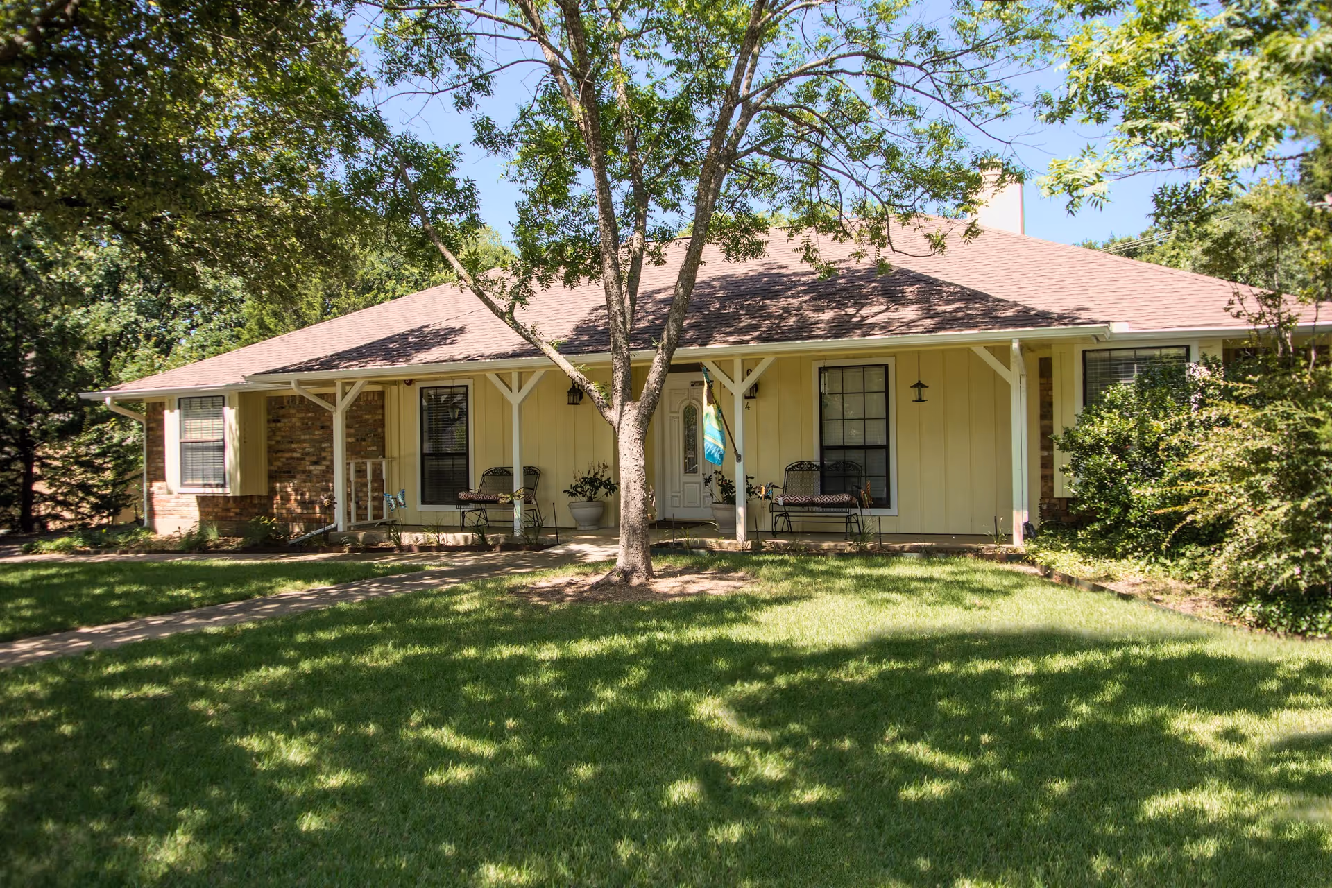 Single-story residential building with a front porch featuring two benches and a potted plant. The building has a light yellow exterior with brick accents and a brown shingled roof. A tree is in the foreground on a well-maintained green lawn, with additional trees and bushes surrounding the building.