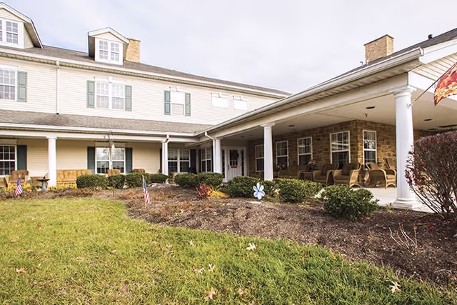 Exterior view of a senior living facility with a covered porch supported by white columns. The building has beige siding with green shutters and multiple windows. There is a landscaped area with bushes, small American flags, and a decorative pinwheel in front of the porch. The sky is overcast.