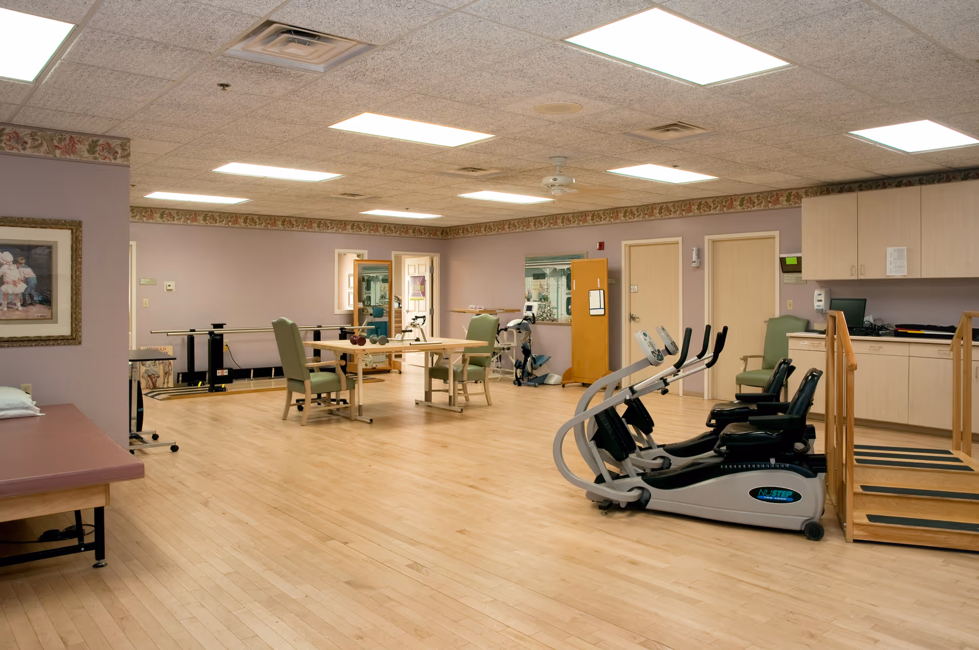 A spacious rehabilitation or physical therapy room with exercise equipment including NuStep machines, parallel bars, tables with chairs, and a wooden ramp. The room has light wood flooring, beige walls with floral border trim, fluorescent ceiling lights, and cabinets along one wall.