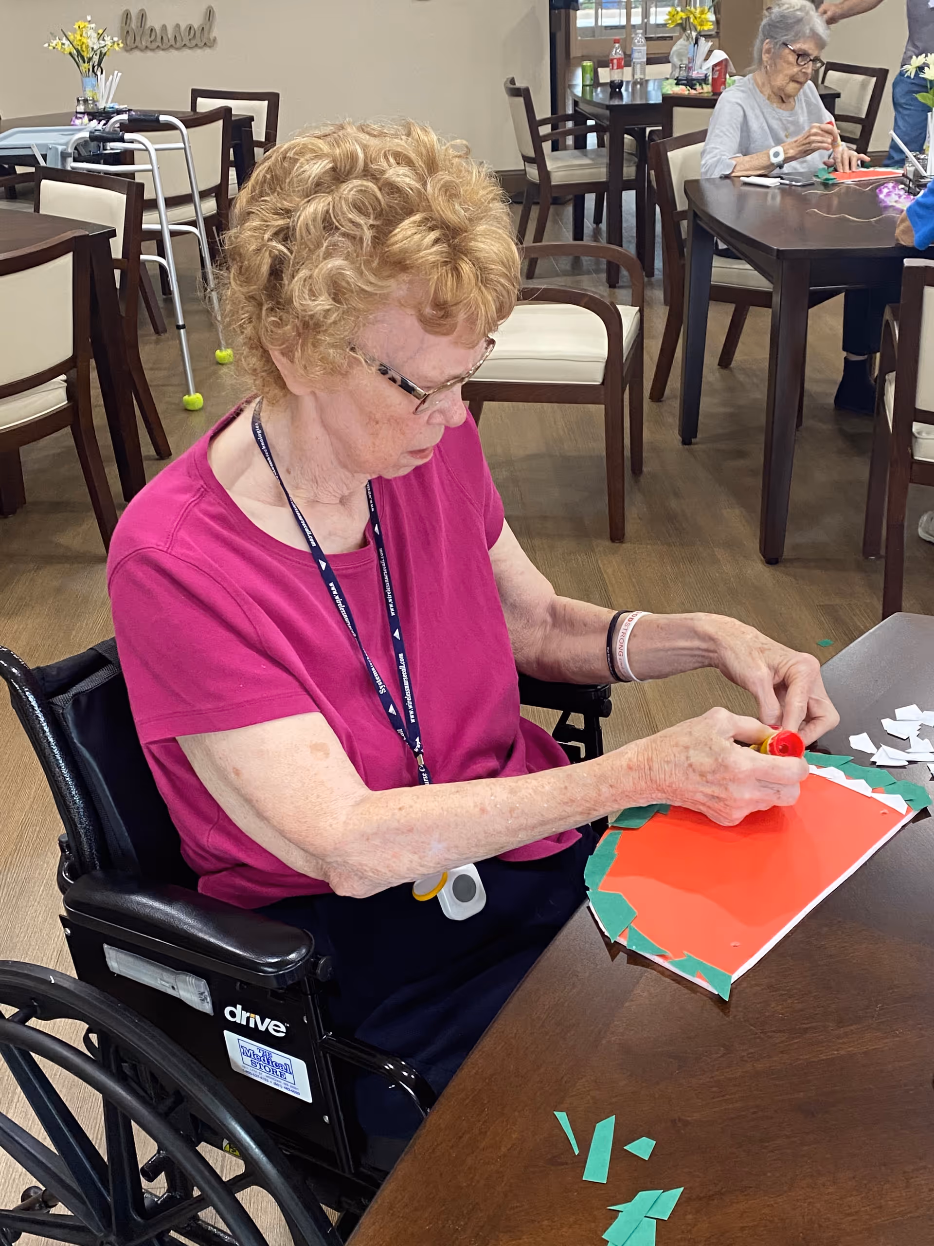 An elderly woman in a wheelchair wearing a magenta shirt is sitting at a table in a communal room, working on a craft project with red, green, and white paper. Other elderly individuals are seated at tables in the background, engaged in similar activities. The room has wooden floors, chairs with cream-colored cushions, and a walker is visible near the wall with a decorative sign that says 'blessed'.