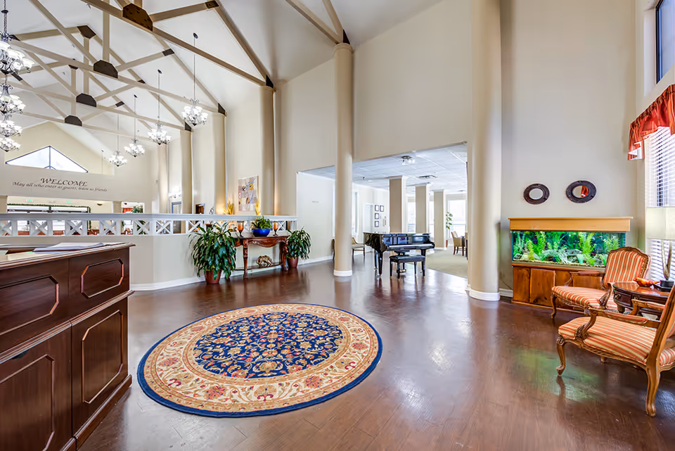 Spacious and well-lit interior of a retirement community featuring a high vaulted ceiling with exposed beams and multiple chandeliers. The room has a polished wooden floor with a round decorative rug in the center. To the right, there is a seating area with two striped armchairs, a side table with a lamp, and a large fish tank. In the background, a black grand piano is visible in an adjacent room with large windows allowing natural light. The reception desk is partially visible on the left side, and there are plants and decorative items along the walls. A welcoming sign is visible on the far wall.