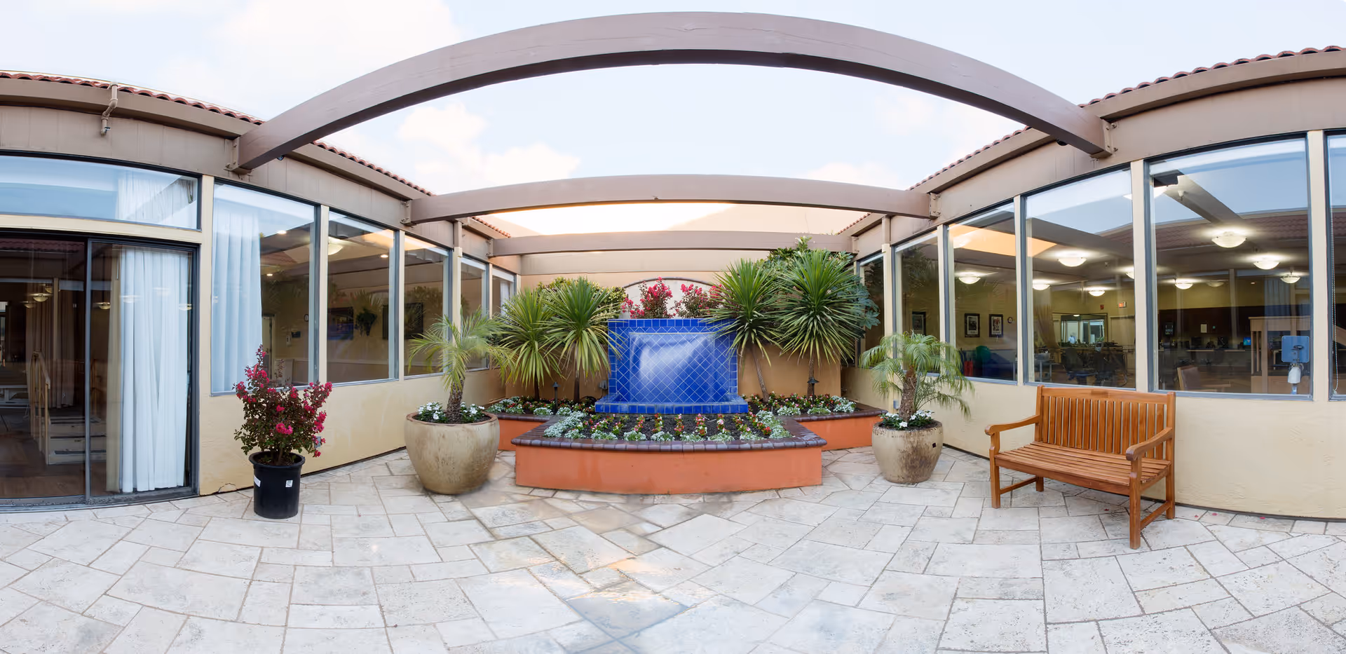 A bright indoor courtyard area with a tiled floor, a blue tiled water fountain surrounded by plants and flowers, large potted plants, and a wooden bench. The courtyard is enclosed by glass windows and doors leading to interior rooms.