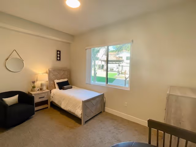 A simple, cozy bedroom with a single bed against the wall, a nightstand with a lamp and small plant, a round mirror hanging on the wall, a black armchair with a pillow, a window showing an outdoor view, and a wooden dresser and chair in the corner.