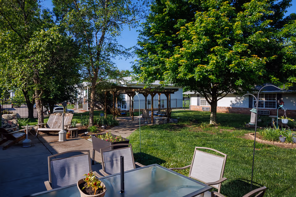 Outdoor patio area at Homestead Assisted Living of Auburn featuring a glass-top table with chairs, wooden benches, a pergola with seating underneath, green grass, trees, and a clear blue sky.
