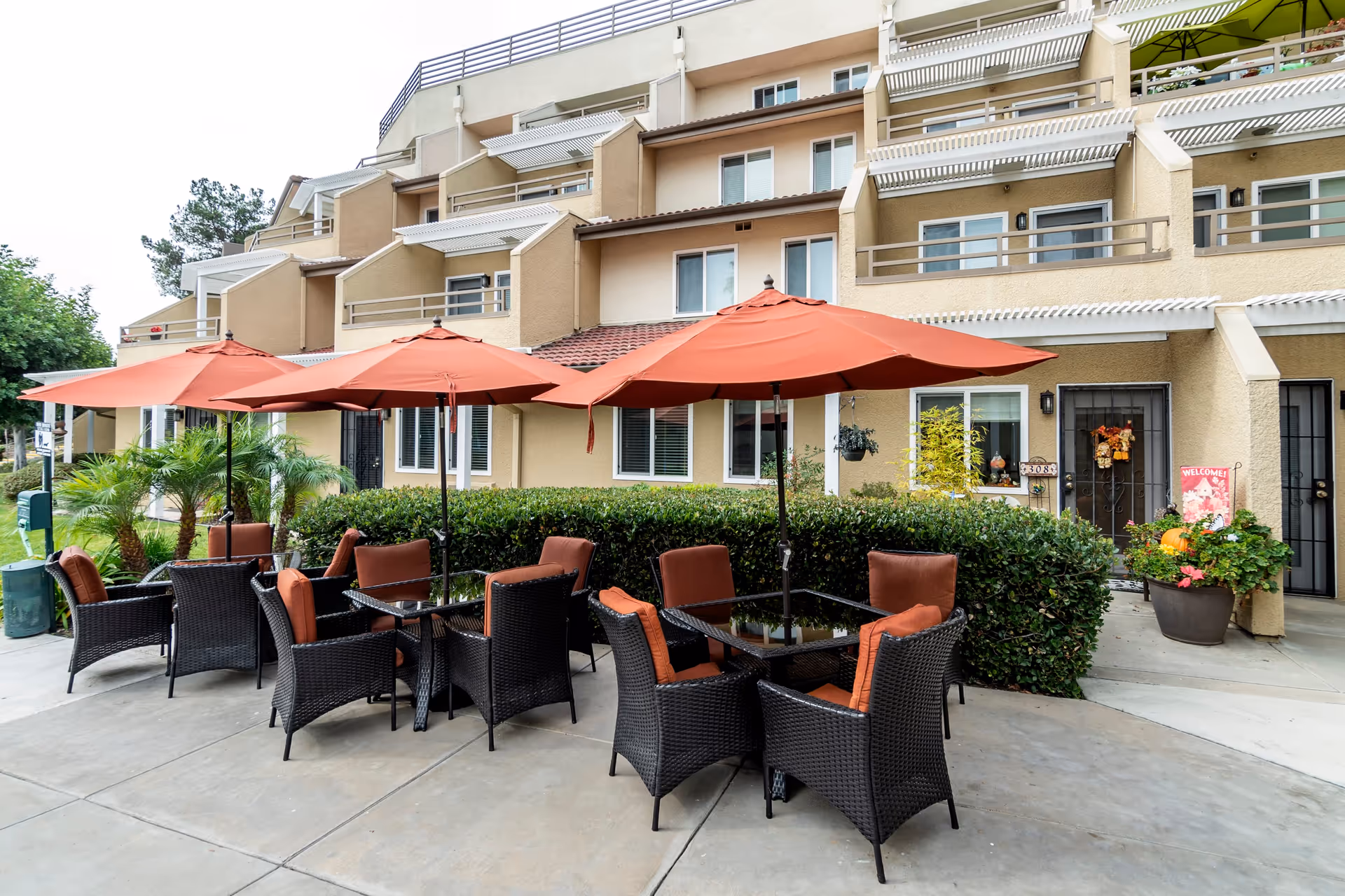 Outdoor patio area at a retirement community with several black wicker tables and chairs, each table shaded by large orange umbrellas. The patio is adjacent to a beige multi-story building with balconies and windows, surrounded by green shrubs and potted plants.