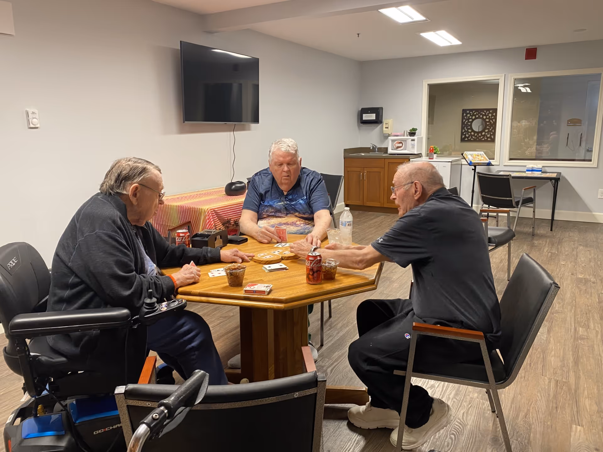 Three elderly men sitting around a wooden table playing cards in a common room with a TV mounted on the wall, a table with a red and yellow striped tablecloth, and a kitchenette area in the background.