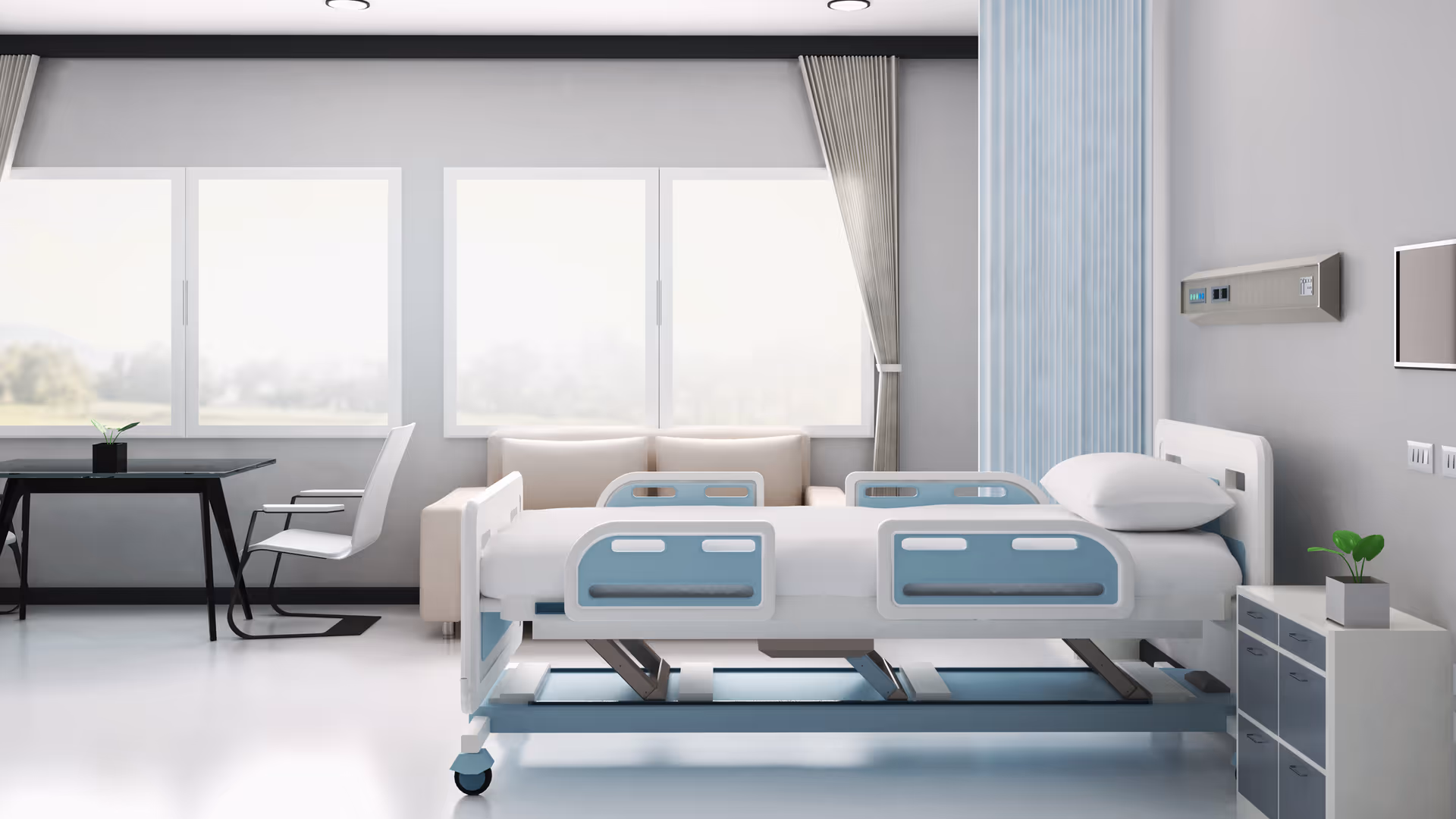 A bright and clean hospital or senior care facility room with a medical bed featuring blue side rails, a white pillow, a small bedside cabinet with a plant, a beige sofa, a black table with a white chair, and large windows with curtains letting in natural light.