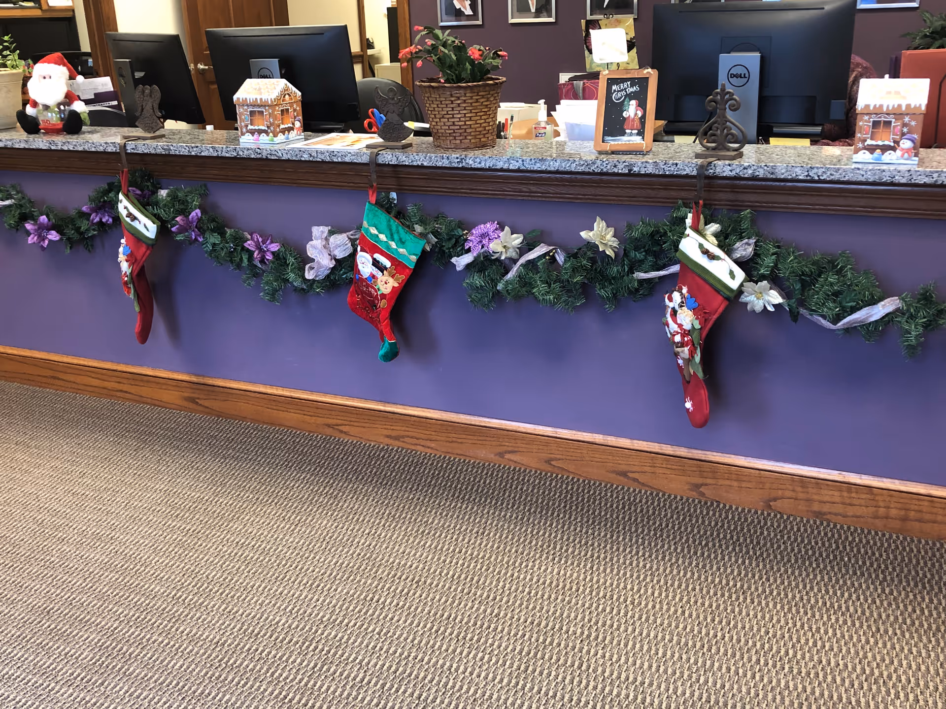 Reception desk decorated with Christmas stockings and garlands with purple flowers. On the desk are holiday decorations including a Santa figurine, a gingerbread house, a potted plant, and a small chalkboard sign that says 'Merry Christmas'. Two computer monitors are visible behind the desk.