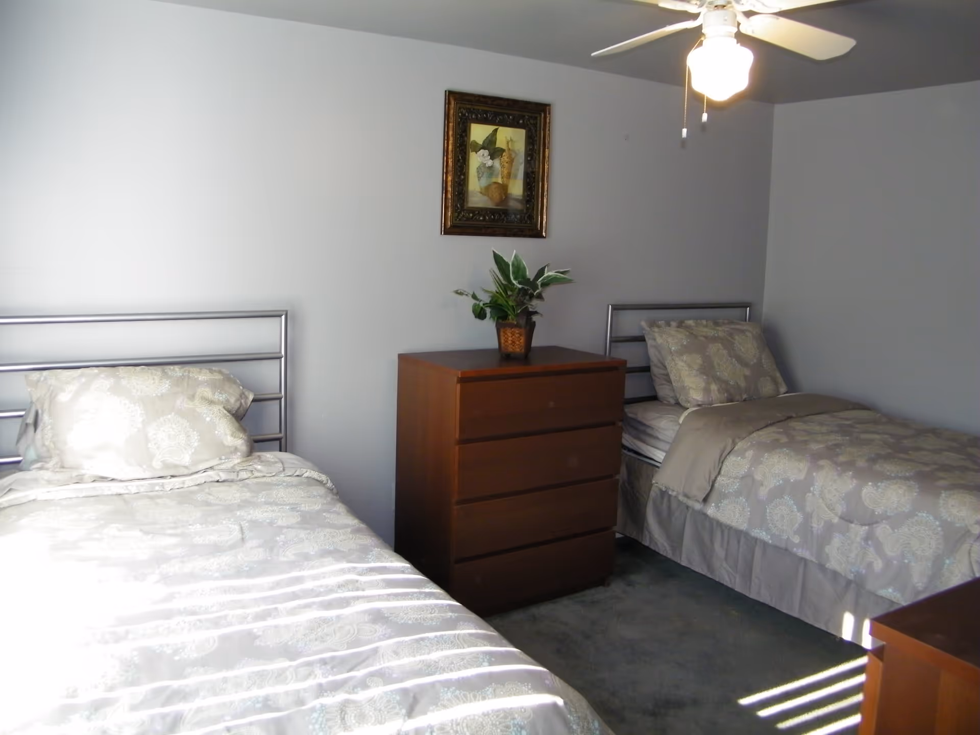 A bedroom with two twin beds featuring matching beige and white patterned bedding. Between the beds is a wooden dresser with a potted plant on top. Above the dresser hangs a framed painting. A ceiling fan with a light is visible on the ceiling.