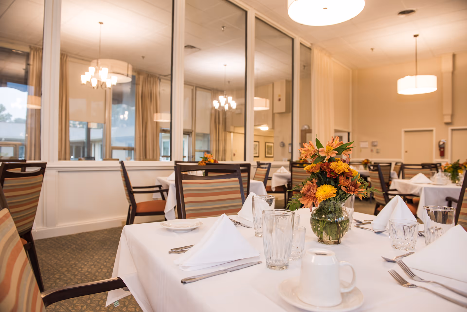 A dining room in a senior living facility with tables covered in white tablecloths, set with folded white napkins, glassware, plates, and silverware. Each table has a vase with a colorful flower arrangement. The room has large windows with beige curtains, carpeted floors, and ceiling lights including chandeliers and pendant lights.