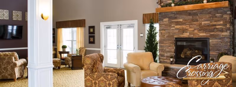 A cozy senior living facility common area with patterned armchairs and a beige armchair arranged around a round leather ottoman. There is a stone fireplace on the right side with a wooden mantel and a small green plant next to it. Double glass doors are visible in the background, along with windows letting in natural light. The room has neutral-colored walls and carpeted floors with a subtle pattern.