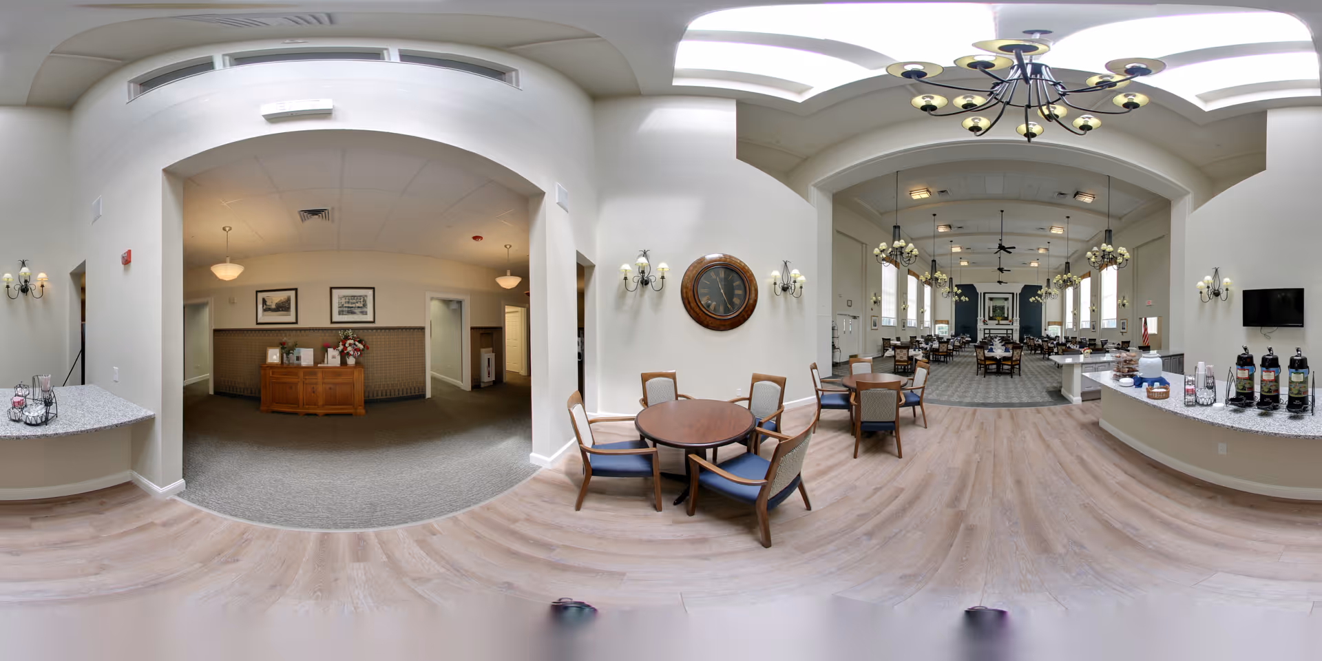 A spacious and well-lit dining area in a senior living facility featuring multiple round wooden tables with chairs, a large clock on the wall, chandeliers hanging from a high ceiling with skylights, and a counter with coffee dispensers and cups. The room has a mix of carpet and wood flooring and an adjacent reception area with a wooden desk and framed pictures on the wall.