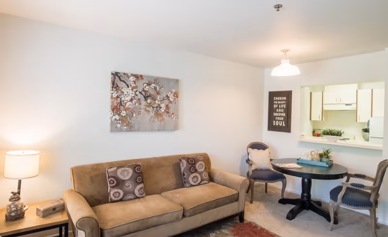 A cozy living room with a beige sofa adorned with two patterned cushions, a side table with a lamp and decorative items, a round black dining table with two upholstered chairs, and a kitchen pass-through window showing white cabinets and a countertop. A floral painting hangs above the sofa, and a framed quote is on the wall near the kitchen opening.
