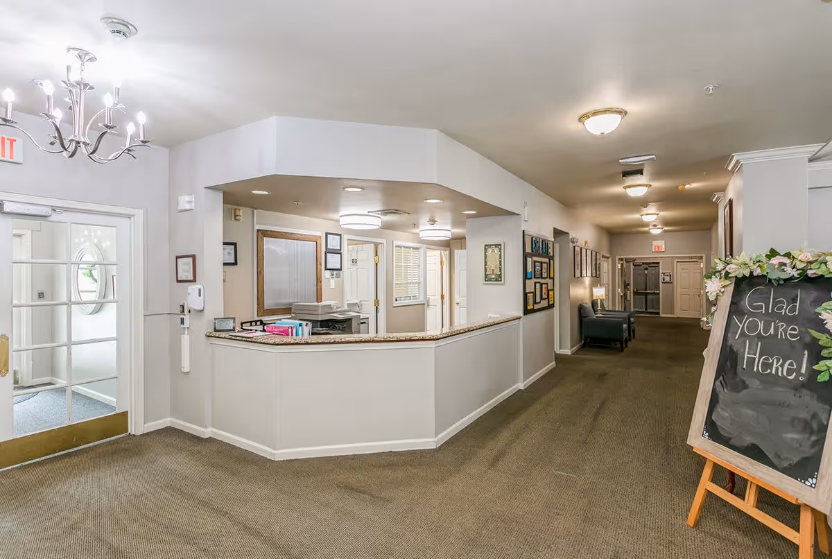 Interior view of a senior living facility reception area with a front desk, a hallway with chairs along the wall, and a chalkboard sign that says 'Glad You're Here!'. The space is well-lit with ceiling lights and a chandelier near the entrance door.