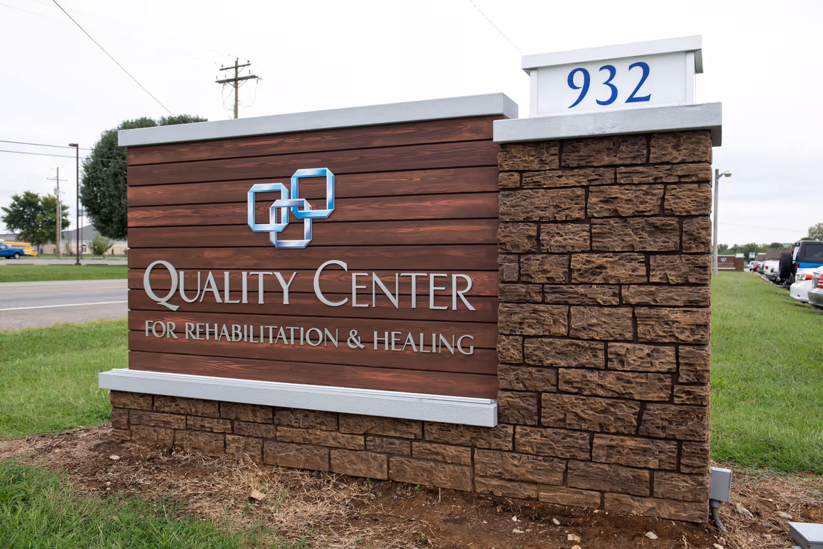 Outdoor sign for Quality Center for Rehabilitation & Healing with address number 932, made of wood and stone materials, located near a road with grass and parked cars visible in the background.