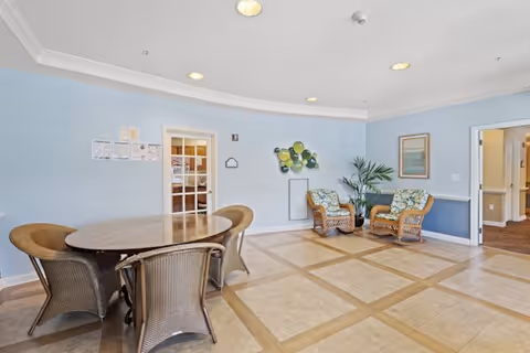 A bright room with light blue walls and tiled floor featuring a round wooden table surrounded by four wicker chairs. In the corner, there are two wicker armchairs with floral cushions and a potted plant. The room has ceiling lights and two doorways leading to other areas.
