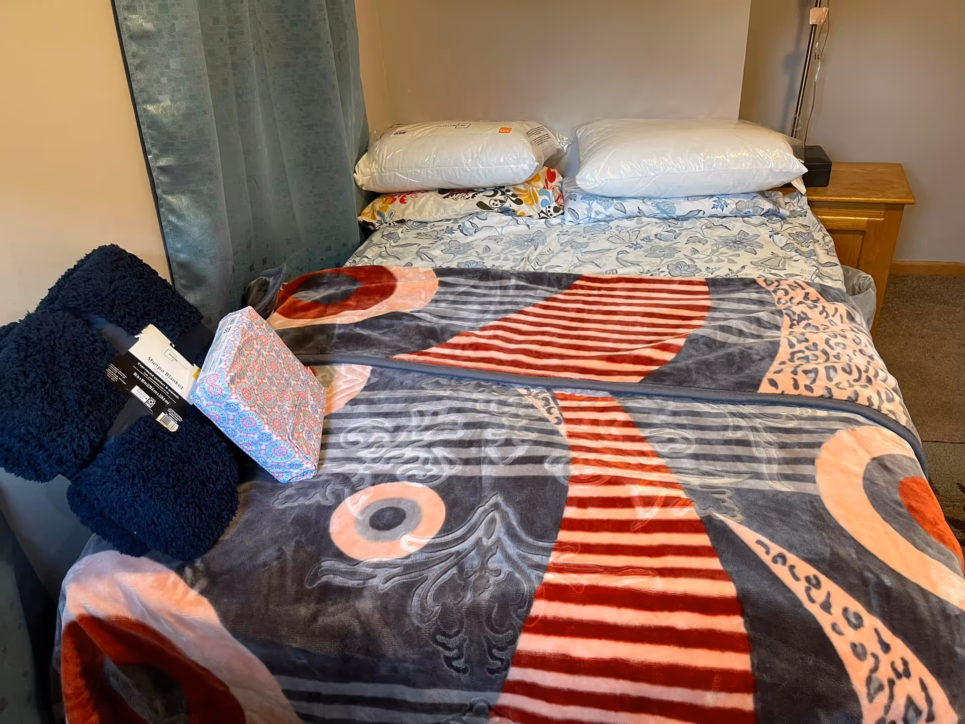 A neatly made bed with two pillows and a colorful patterned blanket in a bedroom. There is a wooden nightstand with a lamp on the right side and a curtain on the left side. Two folded navy blue blankets and a box of tissues are placed on the bed.
