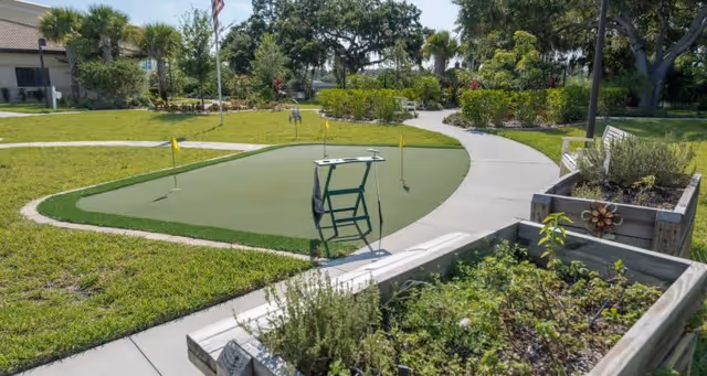 Outdoor putting green with three small flags and a golf ball on the green, surrounded by a curved concrete pathway. Raised garden beds with various plants and a bench are visible on the right side. Trees and a building are in the background under a clear sky.