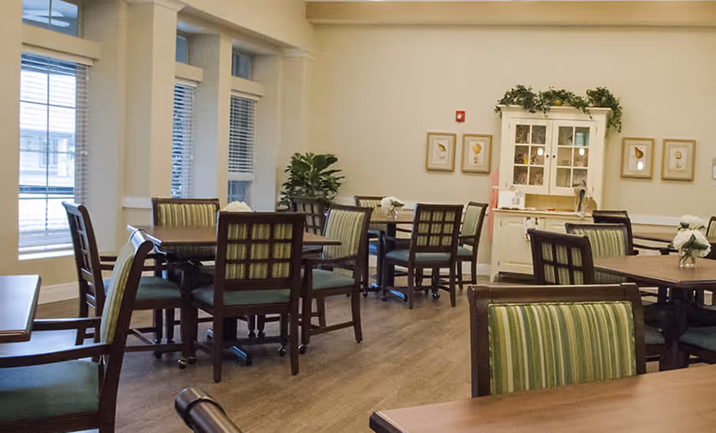 A dining room with several wooden tables and chairs with green striped cushions. The room has large windows with blinds, light-colored walls, a wooden floor, and a white cabinet with glass doors decorated with plants and framed pictures on the wall.