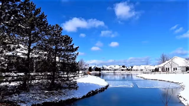 A snowy outdoor scene featuring a partially frozen pond surrounded by snow-covered ground and trees on the left side. In the background, there are white residential buildings under a bright blue sky with scattered clouds.