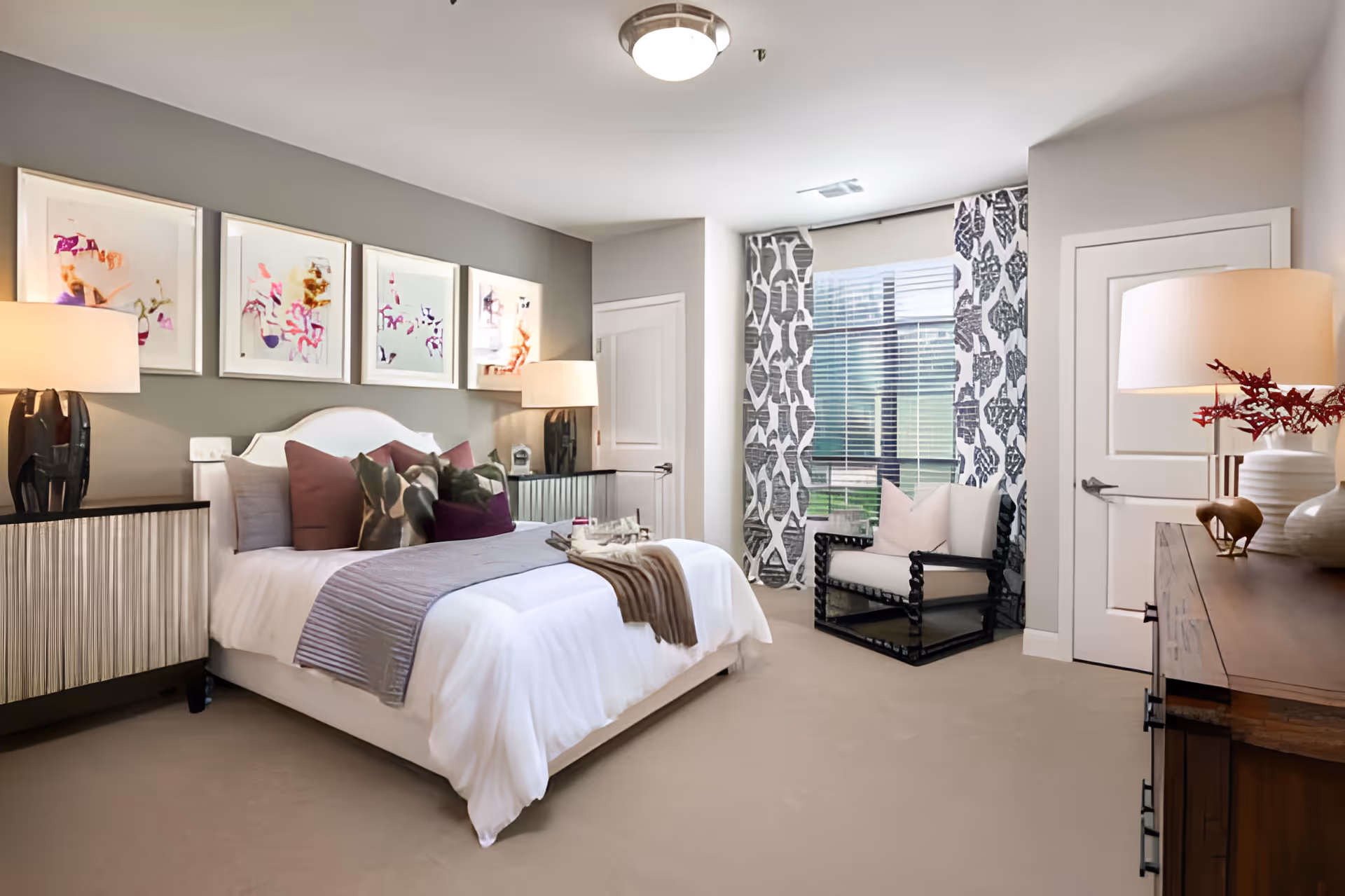 A bright, tastefully decorated bedroom with a made bed, bedside lamps, artwork above the headboard, and an armchair by a window.
