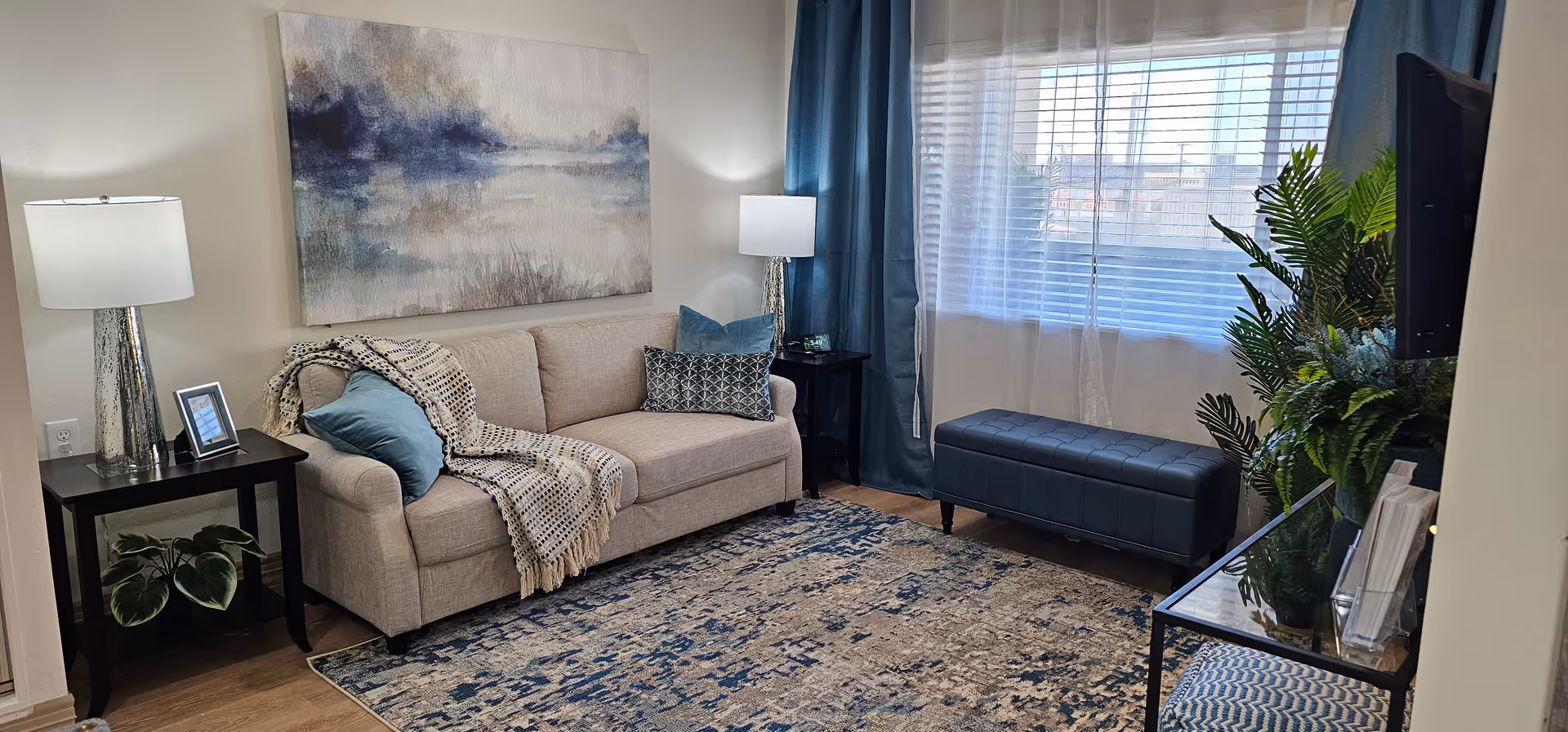 A cozy living room with a beige sofa adorned with blue and patterned pillows and a throw blanket. There are two black side tables with silver lamps on each side of the sofa. A large abstract painting hangs above the sofa. A blue cushioned bench is placed in front of a window with sheer white and blue curtains. A glass-top table with plants and magazines is on the right side, and a TV is mounted on the wall. The room has a blue and beige patterned rug on a wooden floor.