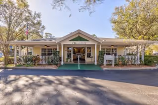 Entrance of a single-story building with a covered porch, white railings, green awning, and surrounding trees.