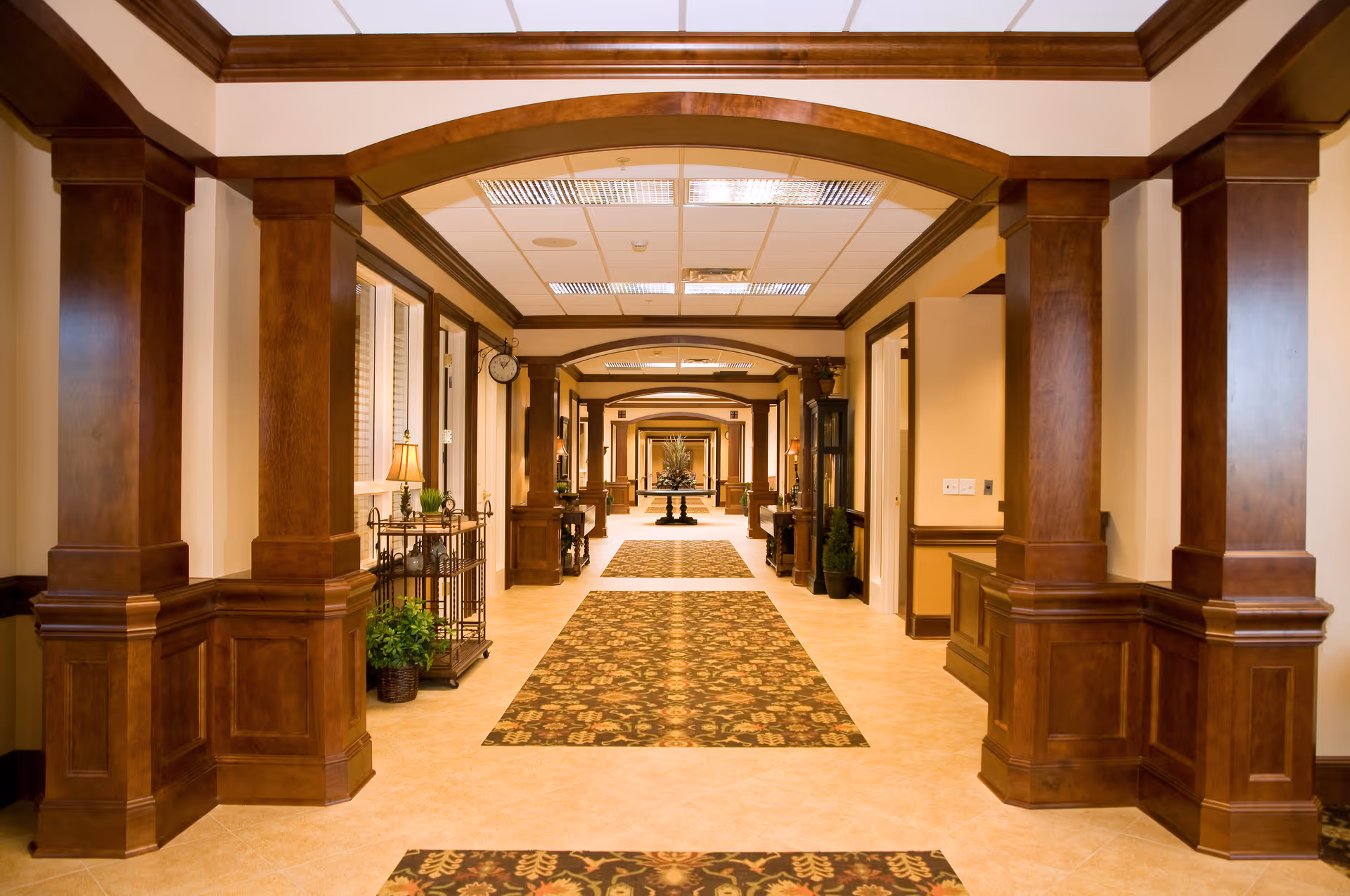 A long, well-lit hallway in a senior living facility with wooden columns and arches, patterned carpet runners on the floor, and decorative plants and furniture along the walls.