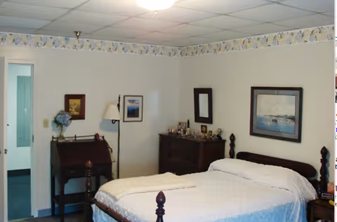 A bedroom with a wooden bed frame and white bedding, a wooden dresser with framed photos and a mirror above it, a small desk with a lamp and a vase of flowers, and framed pictures on the walls. The room has a floral wallpaper border near the ceiling and a door leading to another room.