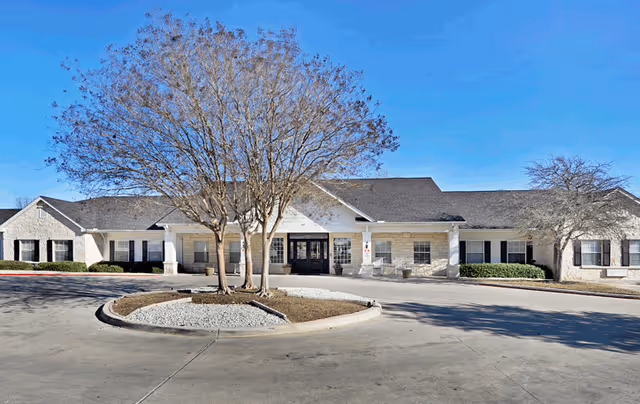 Front exterior of a single-story senior living building with a circular driveway, trees, and a clear blue sky.