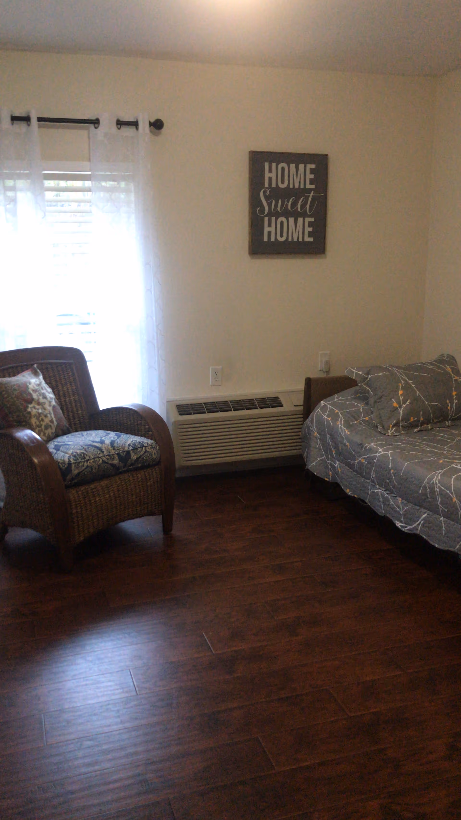 A cozy room with a wicker armchair featuring patterned cushions, a bed with a gray and white patterned comforter, a window with sheer white curtains, and a wall decoration that reads 'HOME Sweet HOME'. The floor is dark wood, and there is a heating/cooling unit beneath the window.