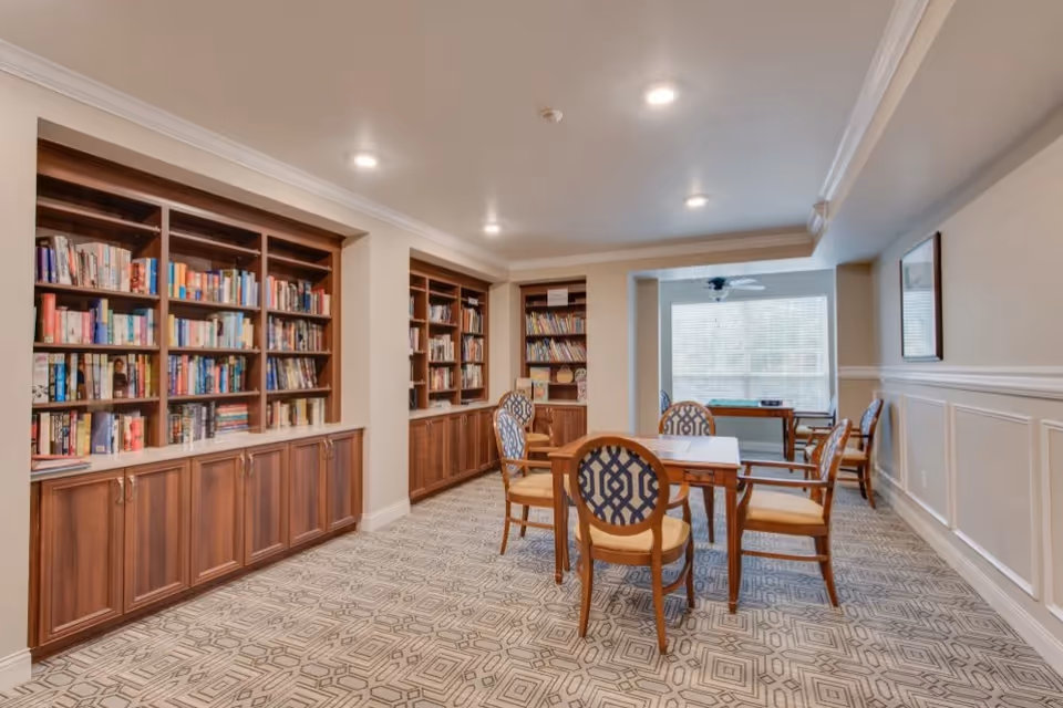 A well-lit room with built-in wooden bookshelves filled with books along the left wall. In the center, there are two wooden tables with six upholstered chairs arranged around them. The room has patterned carpet flooring, white walls with wainscoting, and a large window at the far end letting in natural light.